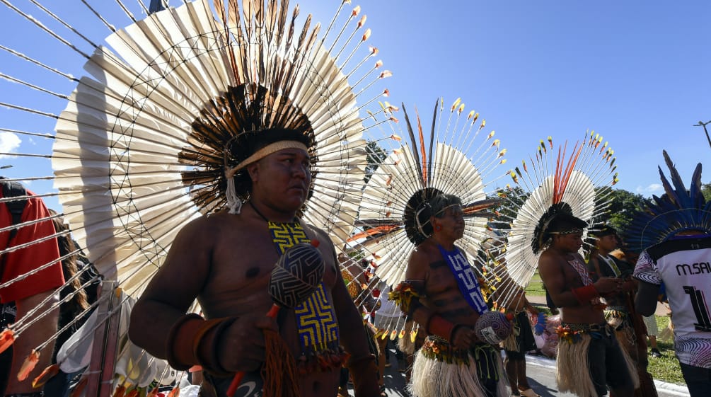 Tre uomini indigeni con grandi copricapi rotondi di piume in testa, con in mano le loro maracas