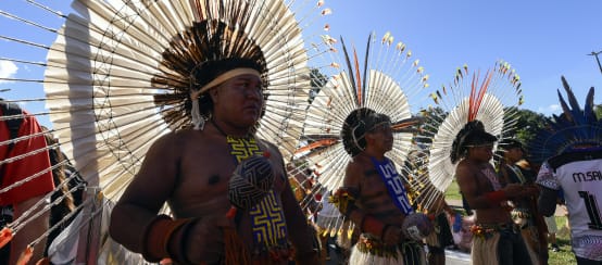 Tre uomini indigeni con grandi copricapi rotondi di piume in testa, con in mano le loro maracas