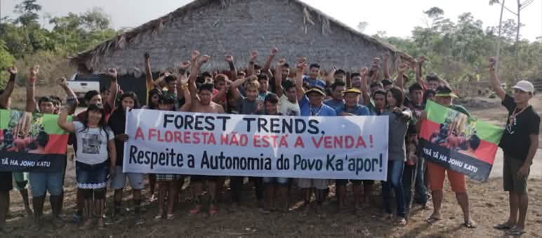 I leader di Tuxa Ta Pame manifestano contro la presenza della ONG Forest Trends nel territorio. Con le braccia tese e i pugni stretti, più di 30 persone di Ka'apor protestano davanti a una casa con pilastri di legno e tetto di paglia. Alcuni di loro reggono uno striscione che recita: "Forest Trends, la foresta non è in vendita".