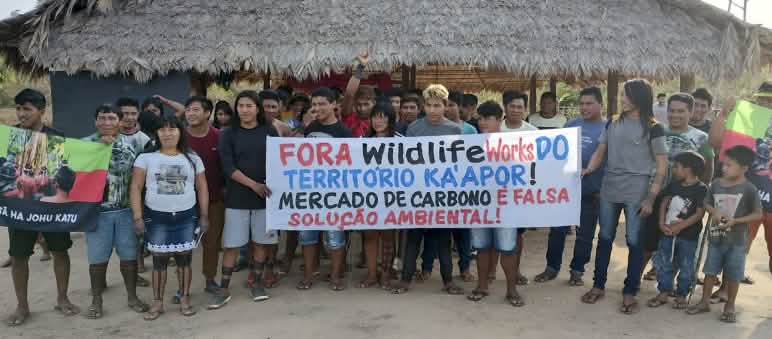 Manifestazione dei leader Ka'apor contro la presenza dell'azienda Wildlife Works nel territorio. Più di 30 indigeni Ka'apor si trovano davanti a una casa tradizionale con pilastri di legno e tetto di paglia. Le persone reggono uno striscione di protesta che recita: "Wildlife Works fuori dal territorio Ka'apor! Il mercato del carbonio è un falso sole