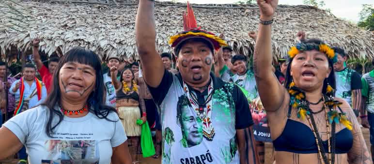Gli indigeni Ka'apor in allerta contro un progetto di commercio di carbonio nel loro territorio Tre indigeni Ka'apor - due donne e un uomo, con i pugni alzati davanti a uno spazio coperto da foglie di palma.