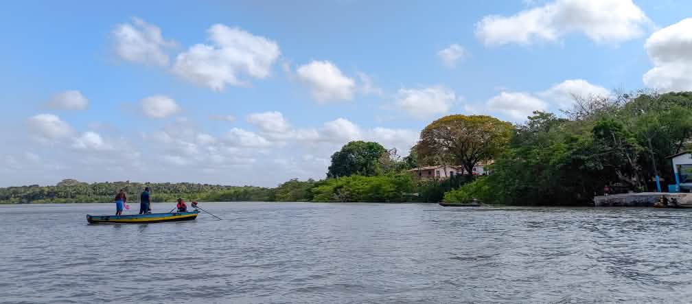 I pescatori artigianali della comunità di Portinho, parte della Resex Tauá-Mirim, mantengono viva la cultura e le conoscenze tradizionali della regione. Sotto un cielo azzurro, tre pescatori su una piccola imbarcazione navigano al largo. A destra si vedono alcune piccole case sulla costa, tra la vegetazione.