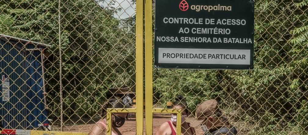 L'azienda agropalma controlla l'accesso della comunità al proprio cimitero. Due donne controllate da una guardia dietro un cancello sbarrato - Testo scritto sul cartello dell'azienda: Agropalma - Controllo dell'accesso al cimitero di Nossa Senhora da Batalha. PROPRIETÀ PRIVATA